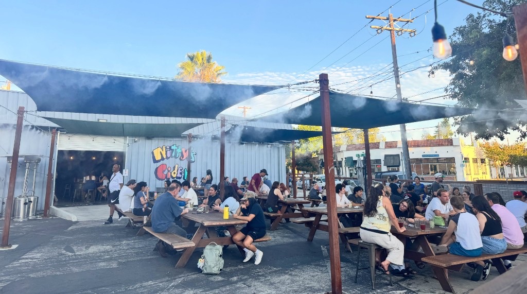 The outdoor patio at Nitty's Cider on a warm evening — friends gathered at picnic tables under shade sails, misters, and string lights, with the colorful Nitty's Cider mural on the wall behind them.